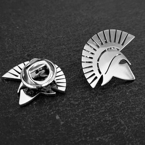 May include: A silver-coloured metal lapel pin shaped like a Spartan helmet with a prominent crest. The pin is shown from two angles, displaying the front face of the helmet and the back side with a circular metal clutch fastener for secure attachment.