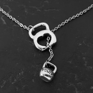 Op de afbeelding: Een zilveren ketting met een hangende kettlebell-bedel. De kettlebell heeft een hart uitgesneden en het woord "POWER" is gegraveerd op de onderkant.