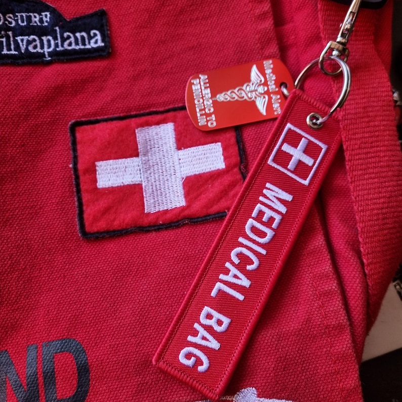 K&ouml;nnte beinhalten: Rote Stofftasche mit einem wei&szlig;en Kreuz-Patch und einem roten und wei&szlig;en medizinischen Beutel-Etikett mit dem Text "MEDICAL BAG" und einem wei&szlig;en Kreuz. Das Etikett ist an einem silbernen Schl&uuml;sselanh&auml;nger mit einem roten medizinischen Warnhinweis befestigt, auf dem "MEDICAL ALERT" und "ALLERGIC TO PENICILLIN" steht.