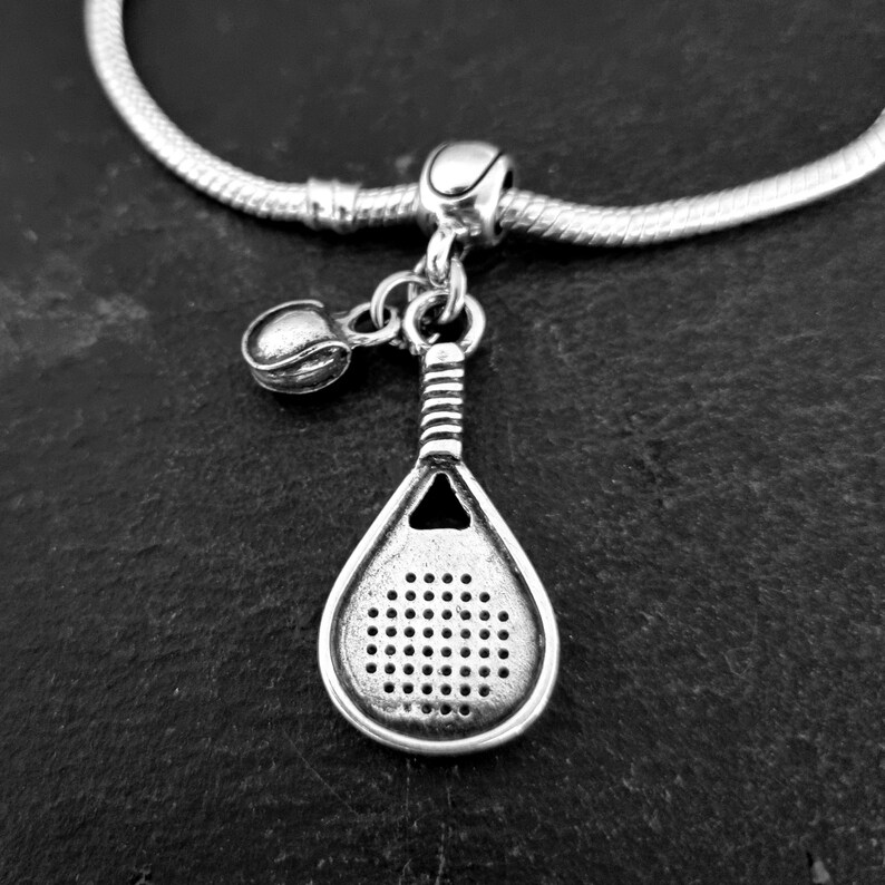 May include: Silver charm bracelet with a paddle tennis racket and ball charm. The charm is detailed with a textured surface and a small hole in the center.