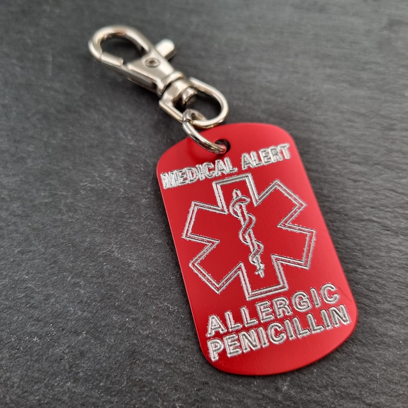 May include: Red metal medical alert tag with a silver star of life symbol and the text "MEDICAL ALERT" and "ALLERGIC PENICILLIN". The tag is attached to a silver keychain.