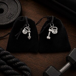 May include: Two small black velvet drawstring pouches, each featuring silver-toned metal charms. One charm is a dumbbell, and the other is a kettlebell engraved with the text WOD & FIT. The pouches are tied with black cords and silver rings.
