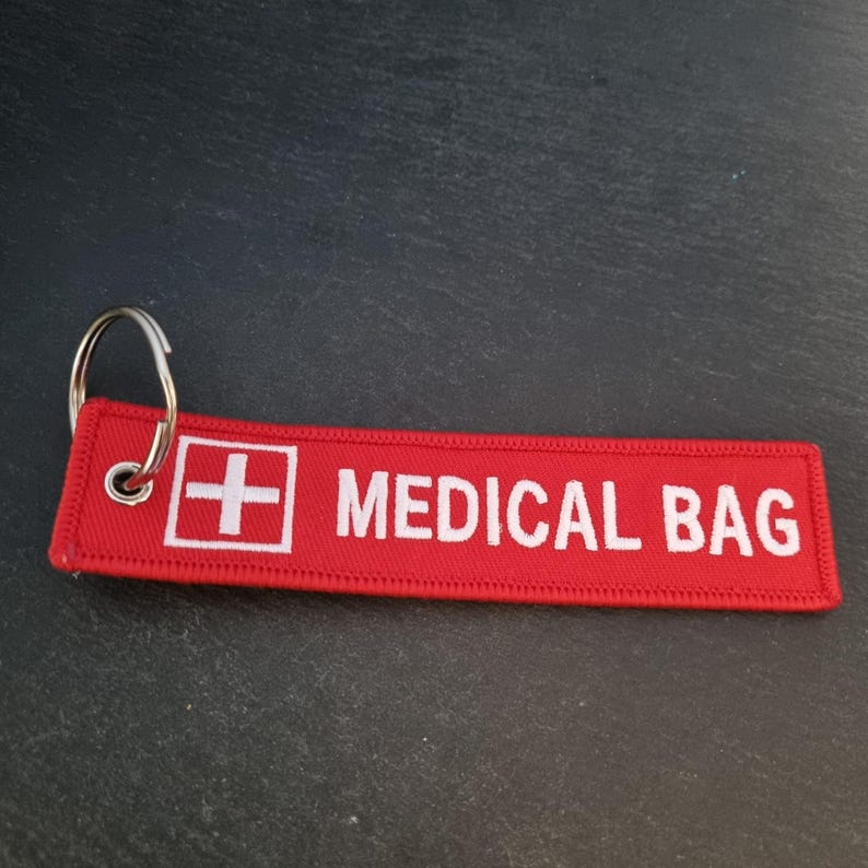 K&ouml;nnte beinhalten: Roter Stoffschl&uuml;sselanh&auml;nger mit einem wei&szlig;en Pluszeichen und dem Text "MEDICAL BAG" in wei&szlig;.