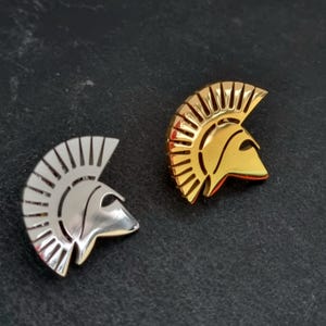 May include: Two metal lapel pins shaped like Spartan helmets, one in a polished silver tone and one in a polished gold tone. Each pin features a high-crested design with vertical ridges and a smooth, curved faceplate, capturing the classic silhouette of an ancient warrior helmet.