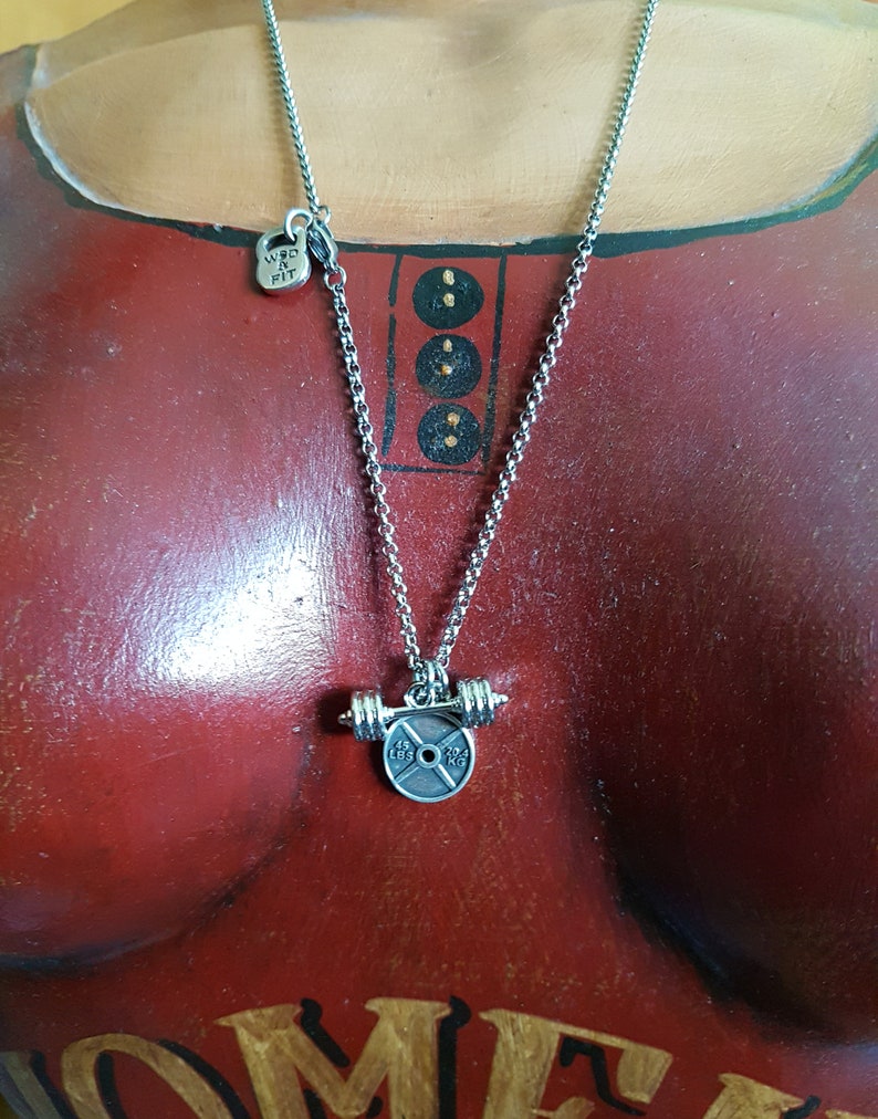 May include: Silver necklace with a weight plate pendant. The plate has the text "15 LBS 7 KG" on it. The necklace also has a small charm with the text "WKD & FIT" on it.