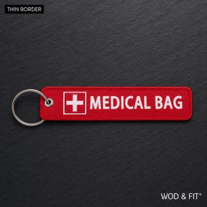 May include: A rectangular red fabric keychain with a white embroidered medical cross symbol and the text MEDICAL BAG in white capital letters. The keychain features a silver metal split ring attached to one end through a reinforced metal eyelet.