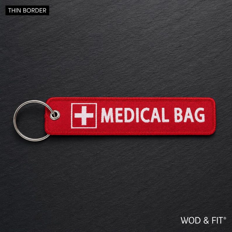 May include: A rectangular red fabric keychain with a white embroidered medical cross symbol and the text MEDICAL BAG in white capital letters. The keychain features a silver metal split ring attached to one end through a reinforced metal eyelet.