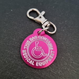 May include: Pink round keyring with a silver clip. The keyring has the text "This bag contains medical equipment" and a wheelchair symbol.