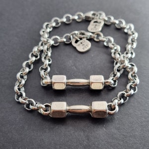 May include: Two silver chain bracelets with dumbbell charms. The charms are silver and have a textured finish. The bracelets are linked together and the charms are hanging from the chains. The text "WRD & FIT" is engraved on one of the charms.