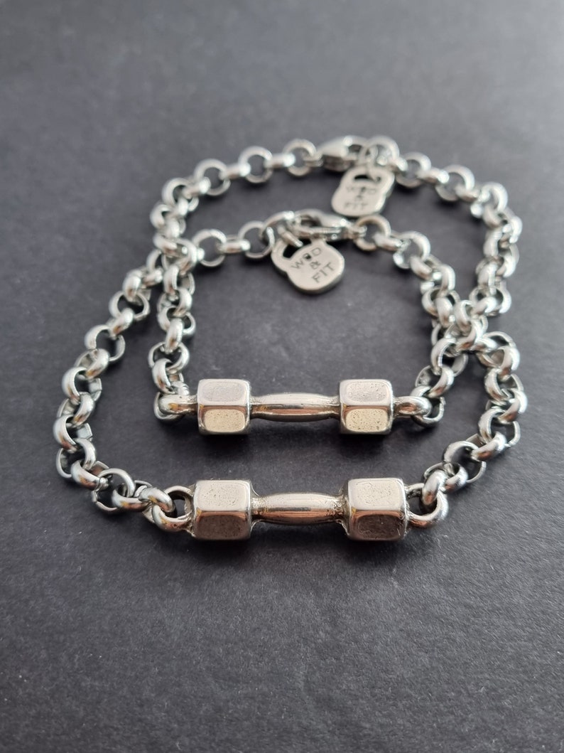 May include: Two silver chain bracelets with dumbbell charms. The charms are silver and have a textured finish. The bracelets are linked together and the charms are hanging from the chains. The text "WRD & FIT" is engraved on one of the charms.