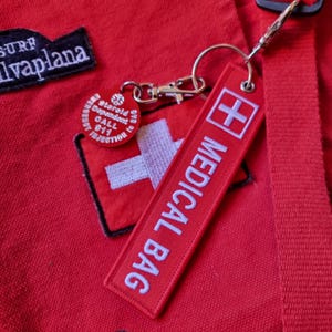 K&ouml;nnte beinhalten: Roter Stoff mit einem wei&szlig;en Kreuz und den Worten "MEDICAL BAG" in Wei&szlig; auf einem roten Stoffanh&auml;nger, der an einem silbernen Schl&uuml;sselband befestigt ist. Der Anh&auml;nger hat ein wei&szlig;es Kreuz auf rotem Hintergrund.