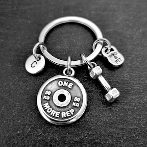 Puede incluir: Llavero plateado con un colgante de disco de pesas que dice "ONE MORE REP". Incluye un dije de mancuerna, un disco pequeño con la letra "C" y un dije que dice "WOD & FIT". El disco de pesas está marcado con "50 KG".