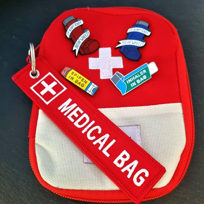 K&ouml;nnte beinhalten: Rote medizinische Tasche mit einem wei&szlig;en Rei&szlig;verschluss und einem wei&szlig;en Canvas-Panel. Die Tasche hat ein wei&szlig; gesticktes Kreuz und den Text "MEDICAL BAG" auf einem roten Riemen. Die Tasche hat mehrere Emaille-Pins, die daran befestigt sind, darunter ein gelber Pin mit dem Text "EPIPEN IN BAG", ein blauer Pin mit dem Text "IT AINT EASY BEIN WHEEZY" und ein roter Pin mit dem Text "IT AINT EASY BEIN WHEEZY".