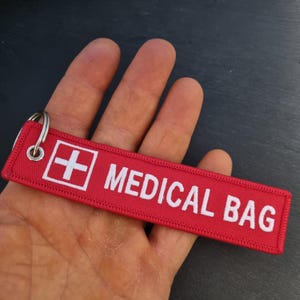 K&ouml;nnte beinhalten: Roter Stoffschl&uuml;sselanh&auml;nger mit wei&szlig;em Text "MEDICAL BAG" und einem wei&szlig;en Pluszeichen in einem roten Quadrat.