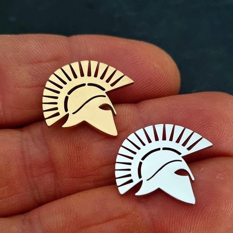 May include: Two small metal pins shaped like Spartan helmets, one in a polished gold tone and the other in a polished silver tone. Each helmet features a distinct crest with vertical cutouts and a pointed visor, showcasing a smooth, reflective surface finish.