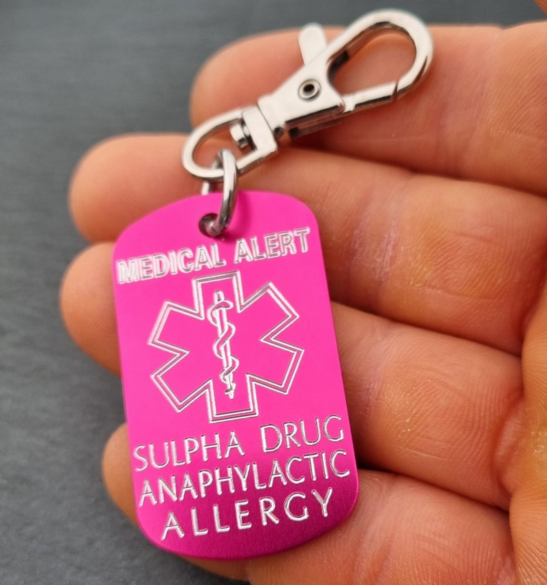 May include: Pink medical alert tag with a silver keychain. The tag has a white star of life symbol and the text "MEDICAL ALERT SULPHA DRUG ANAPHYLACTIC ALLERGY".