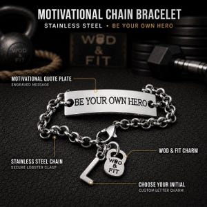 Op de afbeelding: Een roestvrijstalen kettingarmband met een rechthoekige plaat gegraveerd met de tekst BE YOUR OWN HERO. De armband bevat een karabijnsluiting, een aangepaste letterbedel en een kettlebell-vormige bedel met de tekst WOD & FIT.