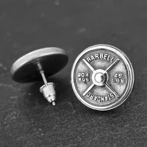 Può includere: Orecchini a bottone a forma di piastra per pesi con bilanciere in argento. Un orecchino è a fuoco, mostrando la parola "BARBELL" e "STANDARD". Il peso è indicato come 20,4 KGS e 45 LBS.