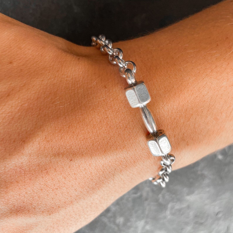May include: A silver chain bracelet with two dumbbell-shaped charms. The bracelet is on a person's wrist.