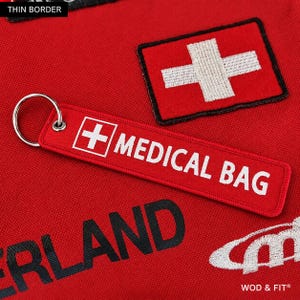 May include: A red rectangular fabric keychain with a silver metal ring. The keychain features a white embroidered Swiss cross and the white text MEDICAL BAG. It is placed on a red fabric surface with a matching embroidered Swiss cross patch.
