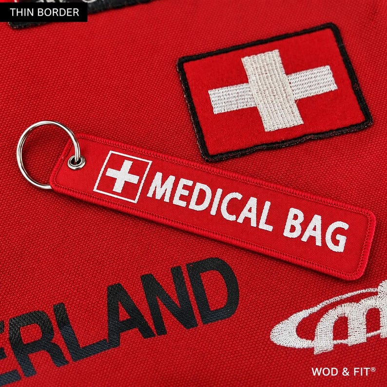 May include: A red rectangular fabric keychain with a silver metal ring. The keychain features a white embroidered Swiss cross and the white text MEDICAL BAG. It is placed on a red fabric surface with a matching embroidered Swiss cross patch.