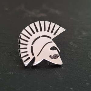 May include: A polished silver-toned metal lapel pin shaped like a Spartan helmet. The design features a high crest with vertical cutouts and a stylised moustache detail on the faceplate, creating a clean, reflective surface with defined lines.