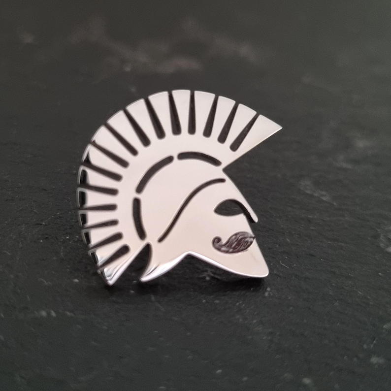 May include: A polished silver-toned metal lapel pin shaped like a Spartan helmet. The design features a high crest with vertical cutouts and a stylised moustache detail on the faceplate, creating a clean, reflective surface with defined lines.