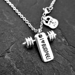 May include: Silver necklace with a dumbbell charm and a tag that says "I love burpees". The necklace also has a small charm shaped like a kettlebell that says "WOD & FIT".