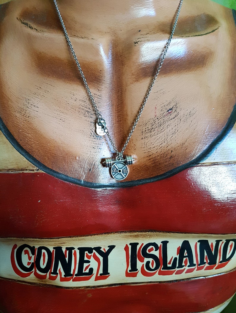 May include: A silver necklace with a barbell weight charm. The charm has a black weight plate with the words "LBS" and "2.5" printed on it. The necklace is on a mannequin wearing a red and white striped shirt with the words "CONEY ISLAND" printed on it.