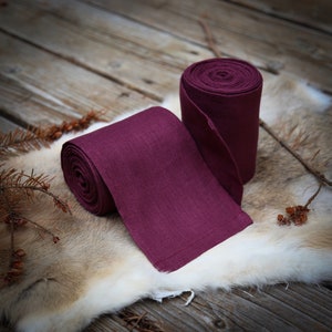 May include: Two rolls of deep burgundy linen ribbon, one unrolled and lying on a light brown fur surface. The ribbon has a subtle texture and a slightly frayed edge.