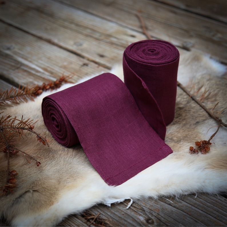 May include: Two rolls of deep burgundy linen ribbon, one unrolled and lying on a light brown fur surface. The ribbon has a subtle texture and a slightly frayed edge.