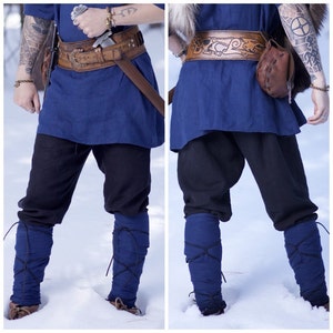 May include: A person wearing a blue tunic with a brown leather belt with a metal buckle and a sword. They are also wearing black pants and blue leg warmers. The person is standing in the snow.