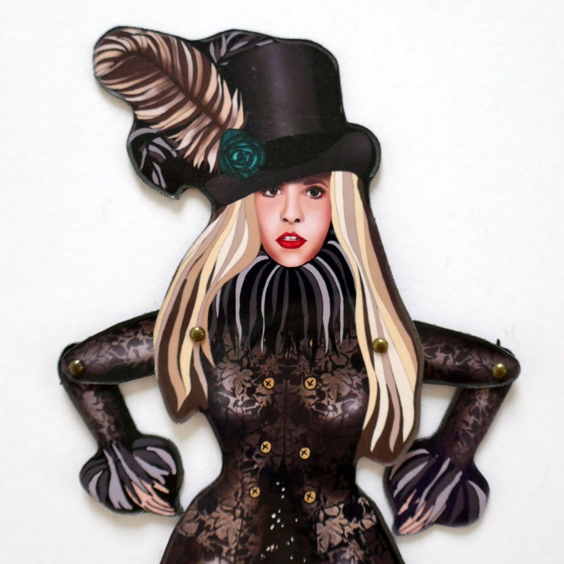 May include: A paper doll with a woman's face, wearing a black top hat with a feather and a teal rose. The doll has long blonde hair and a black lace-patterned bodice. The arms and legs are articulated with brass rivets.
