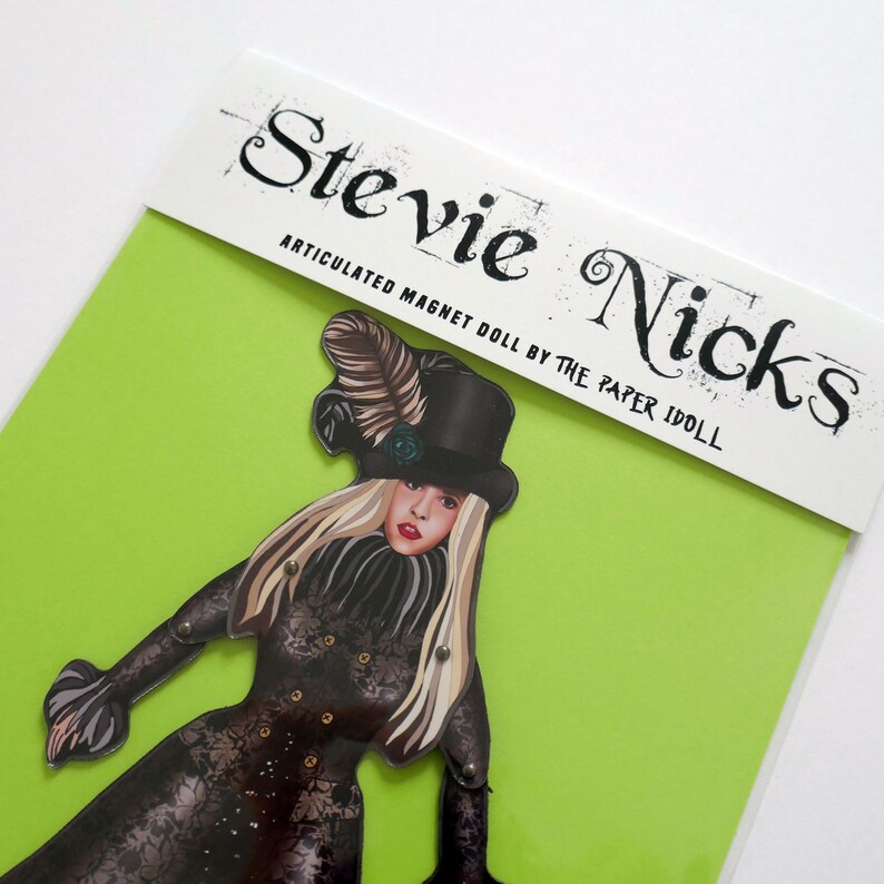 May include: Articulated magnet doll featuring a woman in a black outfit and top hat. The doll has long blonde hair and is on a bright green background. The text "Stevie Nicks" is printed at the top.