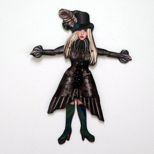 May include: A decorative cutout of a woman in a black Victorian-style dress and top hat. The dress features a floral lace pattern and gold-colored studs. The woman has blonde hair, red lips, and is wearing green boots.
