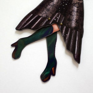 May include: A paper cutout of a figure with a dark floral patterned skirt and green boots. The boots have a high heel and a brown sole. The skirt is held together with two gold-colored buttons. The figure's legs are visible.