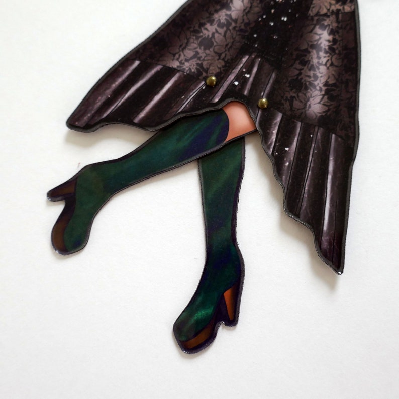 May include: A paper cutout of a figure with a dark floral patterned skirt and green boots. The boots have a high heel and a brown sole. The skirt is held together with two gold-colored buttons. The figure's legs are visible.
