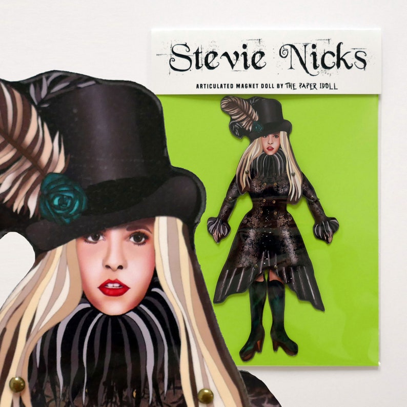 May include: An articulated magnet doll of Stevie Nicks, with a black top hat, long blonde hair, and a black dress. The doll is on a green background with the text "Stevie Nicks" and "ARTICULATED MAGNET DOLL BY THE PAPER IDOLL".