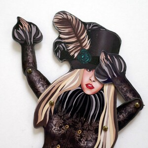 May include: A paper doll figure with a woman's face, wearing a black hat with a feather and a teal rose. The figure has long blonde hair and a black lace bodice. The arms are articulated with brass rivets.