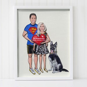 May include: A framed artwork featuring a couple and a dog. The man wears a Superman shirt, and the woman has a floral dress. They hold a heart-shaped sign that reads "1st Anniversary." A German Shepherd sits beside them. The artwork is in a white frame.
