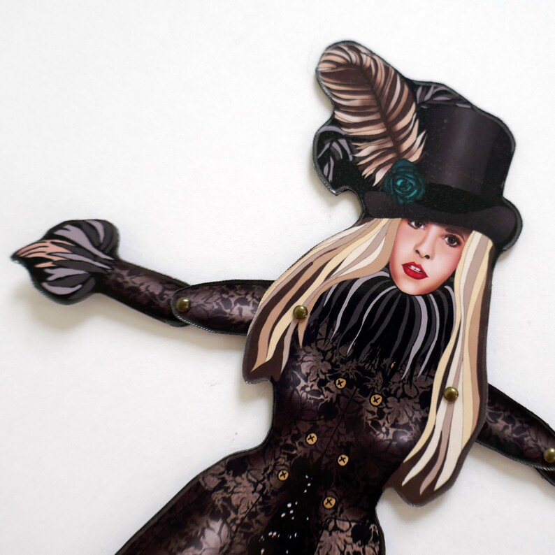 May include: A decorative cutout of a woman wearing a black top hat with a feather and a rose. The cutout has long blonde hair and a black lace dress. The arms and body are jointed with brass fasteners.