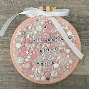 May include: A pink embroidery hoop filled with pink and white beads, pearls, and star-shaped embellishments. The text "A DIAMONDS GOTTA SHINE" is spelled out with letter beads. A white ribbon is attached at the top.