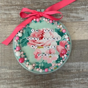 May include: A decorative embroidery hoop with a floral fabric background and the text "I'M THE BEST THING AT THIS PARTY". The hoop is adorned with pink, white, and teal beads and a pink ribbon bow.
