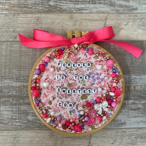 May include: A round embroidery hoop with a floral fabric background. The text "FOREVER IS THE SWEETEST CON" is spelled out with white letter beads. The hoop is decorated with pink and white beads, a pink bow, and a small cowboy boot charm.