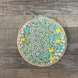 May include: Round embroidery hoop with a floral fabric background. The text "DON'T LET THE HARD DAYS WIN" is spelled out with white letter beads. Embellishments include yellow and blue butterfly shapes and white pearl beads.