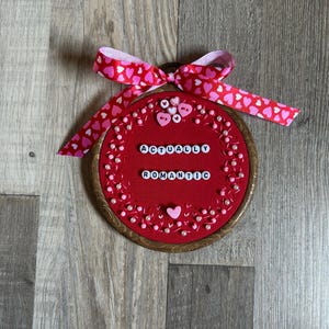 May include: A red embroidery hoop with the words "ACTUALLY ROMANTIC" in white beads. The hoop is decorated with pink hearts, pearls, and a pink and red heart-patterned ribbon. A small pink heart is also present.