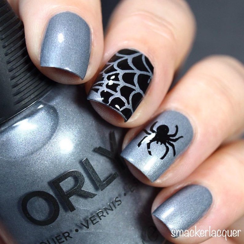 May include: A bottle of gray nail polish with the brand name "ORLY" on the label. The bottle is next to a hand with four fingernails painted in a matching gray color. Two nails have black spider web and spider designs.