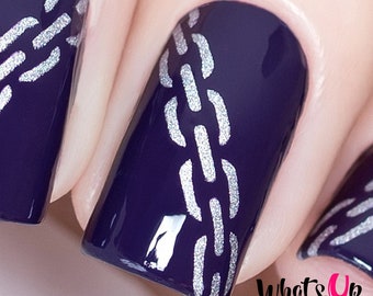 Puede incluir: Primer plano de una manicura de color morado oscuro con un diseño de cadena plateada. El esmalte de uñas es brillante y lustroso. El diseño es intrincado y detallado. "What's Up Nails" está escrito en blanco sobre un fondo rosa.