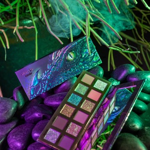 May include: A green and purple eyeshadow palette with a dragon design on the cover. The palette is open and shows 10 different shades of eyeshadow. The palette is sitting on a bed of black and purple stones.