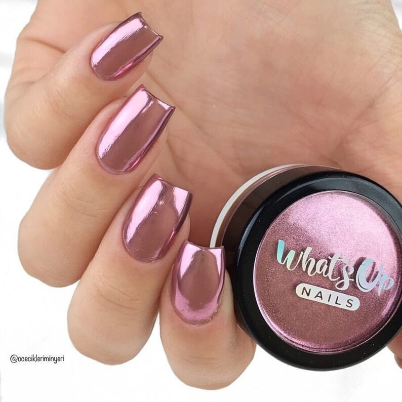 May include: A close-up of a jar of pink chrome nail powder. The jar has a black lid with the text "What's Up Nails" printed on it. The powder is a fine, shimmery texture and is perfect for creating a holographic effect on nails.